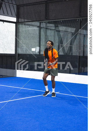 African American man standing on blue padel court holding racket wearing orange shirt olive shorts African American man standing on blue padel court holding racket wearing orange shirt olive shorts 140927766