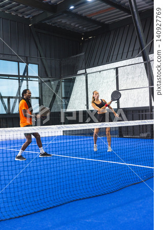 Diverse padel partners standing on bright blue court under metal roof holding rackets by fencing Diverse padel partners standing on bright blue court under metal roof holding rackets by fencing 140927769