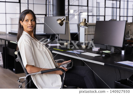 Asian woman sitting in swivel chair at open-plan office holding tablet, turning gaze right Asian woman sitting in swivel chair at open-plan office holding tablet, turning gaze right 140927930