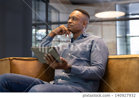 African american man wearing blue shirt sitting in office holding tablet, wearing smartwatch African american man wearing blue shirt sitting in office holding tablet, wearing smartwatch 140927932