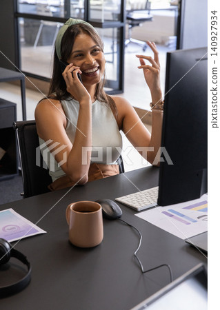 Woman speaking on smartphone, gesturing at desk in open-plan office with printed charts 140927934