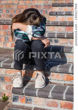 Teenteenage girl sitting on brick steps in plaid shirt hugging knees leaning against brick wall Teenteenage girl sitting on brick steps in plaid shirt hugging knees leaning against brick wall 140927948
