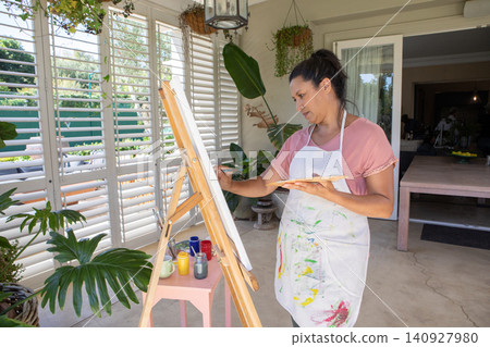 Woman painting on canvas on veranda wearing paint-stained apron, holding palette, copy space Woman painting on canvas on veranda wearing paint-stained apron, holding palette, copy space 140927980