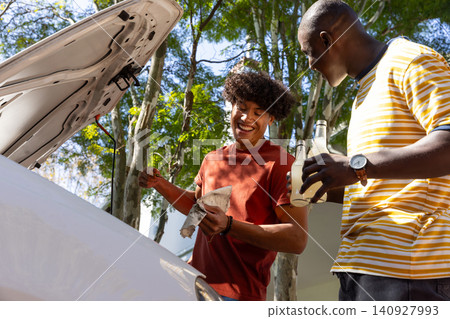 Diverse male friends checking oil dipstick under car hood with bottled drinks on driveway Diverse male friends checking oil dipstick under car hood with bottled drinks on driveway 140927993