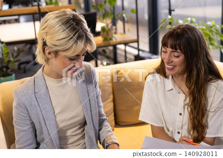 Female colleagues reviewing document with pen on mustard sofa in office lounge Female colleagues reviewing document with pen on mustard sofa in office lounge 140928011