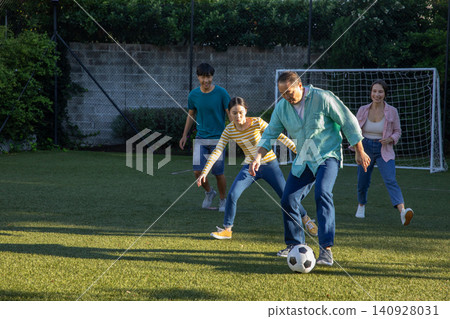 Diverse family playing soccer on grass field next to brick wall with soccer ball and goal net Diverse family playing soccer on grass field next to brick wall with soccer ball and goal net 140928031