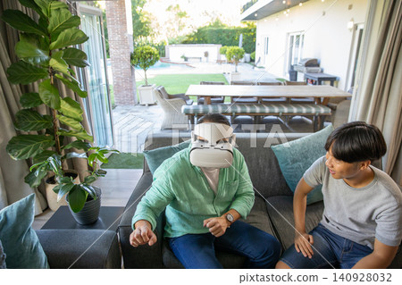Asian father using vr headset while daughter watching on sofa by glass doors to patio Asian father using vr headset while daughter watching on sofa by glass doors to patio 140928032