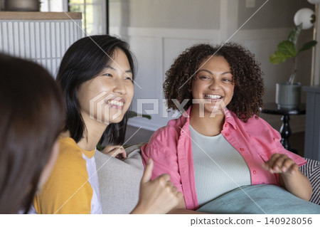 Diverse female friends laughing and chatting on light-colored sofa at home by side table with plant Diverse female friends laughing and chatting on light-colored sofa at home by side table with plant 140928056