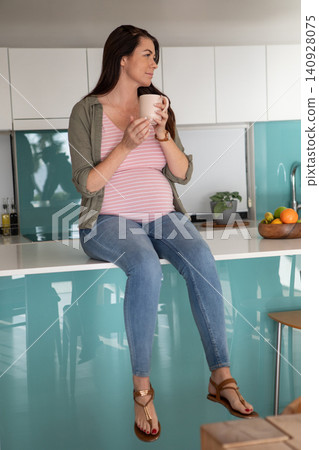 Mid adult pregnant woman sitting on kitchen island holding mug beside fruit bowl while gazing left 140928075