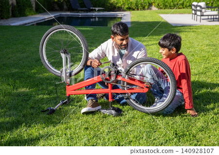 Asian father with son kneeling on backyard lawn by pool, repairing bright red bicycle with wrench 140928107