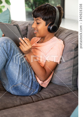 South asian school-age boy sitting on gray sofa in living room with tablet wearing headphones South asian school-age boy sitting on gray sofa in living room with tablet wearing headphones 140928108