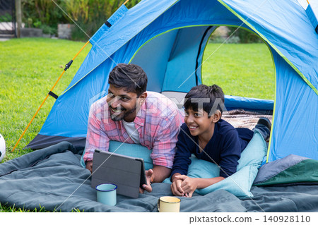 South asian father and son lying on camping blanket in front of blue tent, using tablet 140928110