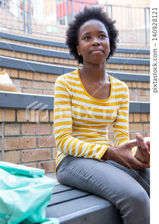 Adolescent african american female student sitting on bench at campus quad next to backpack Adolescent african american female student sitting on bench at campus quad next to backpack 140928121