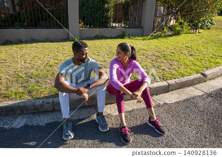 Diverse workout partners sitting on curb by road talking, in running shoes and resting Diverse workout partners sitting on curb by road talking, in running shoes and resting 140928136