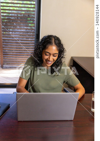 Indian woman smiling while typing on laptop at wooden desk with notebook and water glass Indian woman smiling while typing on laptop at wooden desk with notebook and water glass 140928144