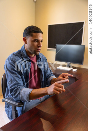 Hispanic man wearing denim shirt sitting at office desk gesturing near monitor, copy space Hispanic man wearing denim shirt sitting at office desk gesturing near monitor, copy space 140928146