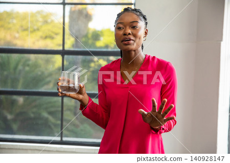 African american woman standing in office by metal window holding clear glass and speaking African american woman standing in office by metal window holding clear glass and speaking 140928147