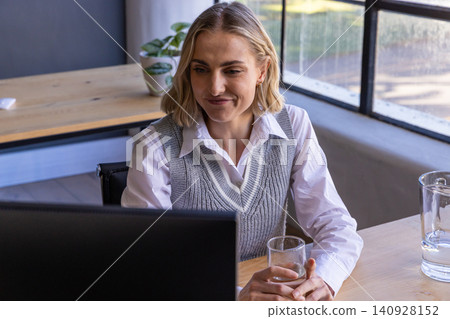 Woman wearing white shirt holding glass of water while sitting at office desk with monitor Woman wearing white shirt holding glass of water while sitting at office desk with monitor 140928152