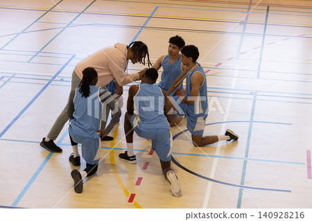 Diverse male basketball coach kneeling in beige hoodie with players in navy blue uniforms on court Diverse male basketball coach kneeling in beige hoodie with players in navy blue uniforms on court 140928216