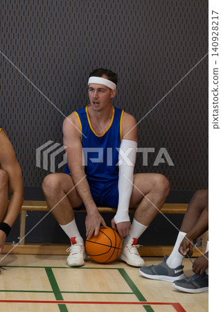 Diverse male basketball teammates in jerseys sitting on gym bench holding orange basketball Diverse male basketball teammates in jerseys sitting on gym bench holding orange basketball 140928217