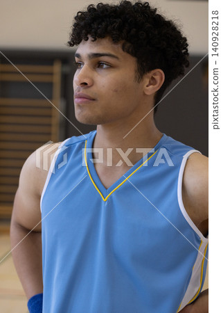 Male athlete standing in gym on basketball court floor beside wooden wall bars wearing blue jersey Male athlete standing in gym on basketball court floor beside wooden wall bars wearing blue jersey 140928218