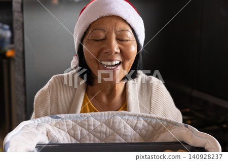 Senior Asian woman leaning over baby bassinet with quilted blanket in living room wearing Santa hat Senior Asian woman leaning over baby bassinet with quilted blanket in living room wearing Santa hat 140928237