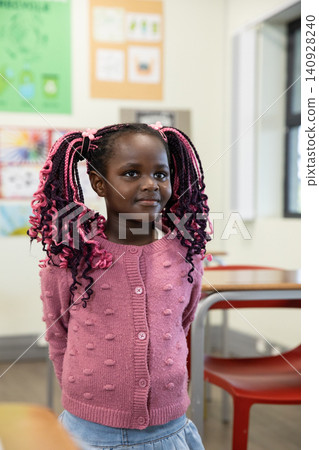 African american girl standing in pink cardigan and skirt in classroom near desks and posters 140928240