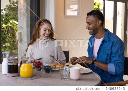 Diverse couple cutting into stack of pancakes and sitting at breakfast nook table with fruit bowls 140928243