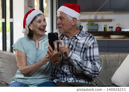 Senior couple wearing Santa hats laughing and pointing at smartphone while sitting on sofa at home Senior couple wearing Santa hats laughing and pointing at smartphone while sitting on sofa at home 140928251