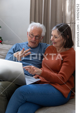 Senior couple sitting on sofa using silver laptop in living room with Christmas tree and curtains Senior couple sitting on sofa using silver laptop in living room with Christmas tree and curtains 140928252