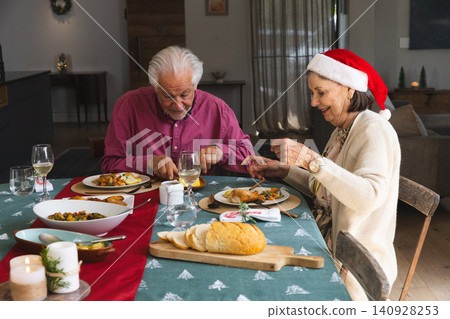 Senior couple wearing Santa hat cutting chicken and vegetables in dining room with wine glasses Senior couple wearing Santa hat cutting chicken and vegetables in dining room with wine glasses 140928253