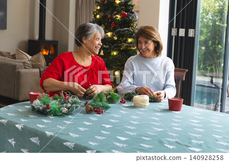 Senior, middle-aged women friends crafting festive wreaths in living room with pine cones and twine Senior, middle-aged women friends crafting festive wreaths in living room with pine cones and twine 140928258