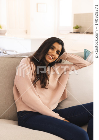 South Asian woman sitting at home on gray sofa wearing pink sweater showcasing kitchen countertop South Asian woman sitting at home on gray sofa wearing pink sweater showcasing kitchen countertop 140928271