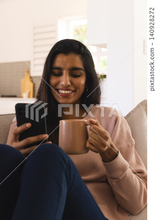Asian woman sitting on couch holding smartphone and ceramic mug near kitchen counter Asian woman sitting on couch holding smartphone and ceramic mug near kitchen counter 140928272