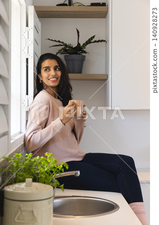 Asian woman holding ceramic mug, sitting on countertop in kitchen by window near sink Asian woman holding ceramic mug, sitting on countertop in kitchen by window near sink 140928273