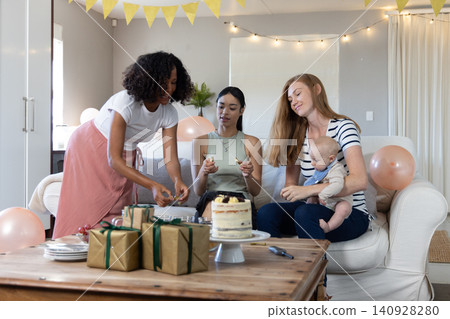 Diverse female friends with infant gathering around sofa at home displaying cake, gifts Diverse female friends with infant gathering around sofa at home displaying cake, gifts 140928280