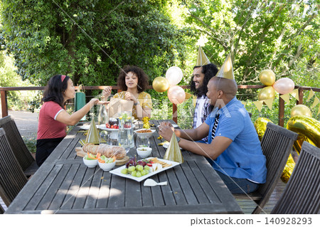 Diverse friends sitting around table on deck sharing gift bag, sampling pastries amid balloons Diverse friends sitting around table on deck sharing gift bag, sampling pastries amid balloons 140928293