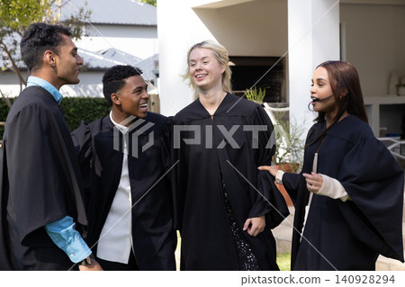 Diverse friends chatting in sunlit yard while wearing black graduation gowns, headset microphone 140928294