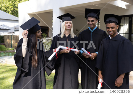 Diverse graduates wearing mortarboard caps, gowns holding diplomas with ribbons on grassy lawn Diverse graduates wearing mortarboard caps, gowns holding diplomas with ribbons on grassy lawn 140928296
