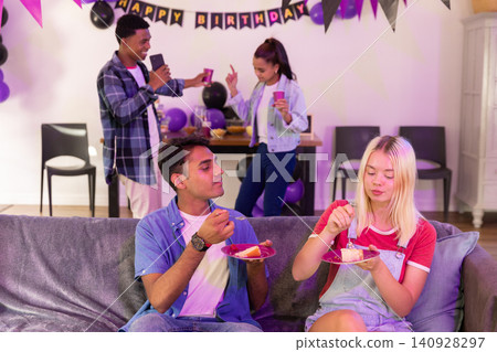 Diverse friends celebrating birthday on grey couch with banner balloons holding cake, pink cups Diverse friends celebrating birthday on grey couch with banner balloons holding cake, pink cups 140928297