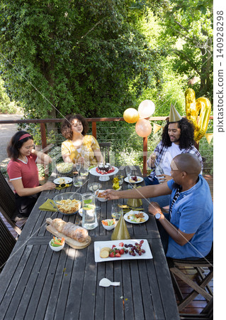 Diverse friends sitting at wooden table on backyard deck, cutting cake, pouring lemon water 140928298