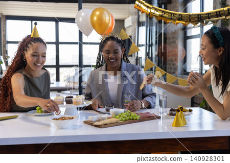 Diverse female coworkers gathering around counter in kitchen serving cupcakes near birthday banner Diverse female coworkers gathering around counter in kitchen serving cupcakes near birthday banner 140928301
