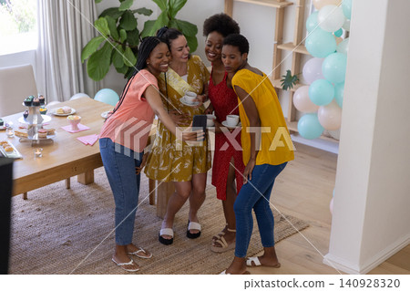Diverse female friends taking group selfie in living room holding coffee cups beside dessert stand Diverse female friends taking group selfie in living room holding coffee cups beside dessert stand 140928320