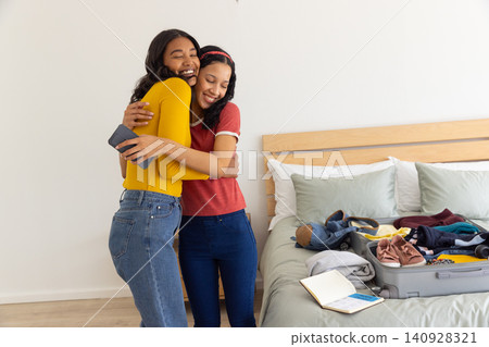 Diverse mom and teenage daughter hugging in bedroom near open suitcase while holding smartphone 140928321