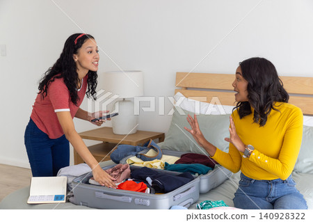 Diverse mom and teenage daughter packing open suitcase on bed using smartphone and notebook Diverse mom and teenage daughter packing open suitcase on bed using smartphone and notebook 140928322