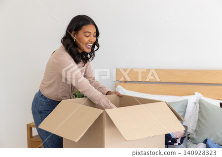 Woman leaning over cardboard box on bed unpacking clothes near plant, copy space Woman leaning over cardboard box on bed unpacking clothes near plant, copy space 140928323