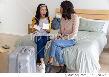 Diverse mom and teenage daughter sitting on bed opening white gift box in bedroom with suitcase Diverse mom and teenage daughter sitting on bed opening white gift box in bedroom with suitcase 140928324