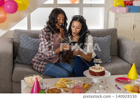 Diverse mom and teenage daughter looking at smartphone at home with birthday cake Diverse mom and teenage daughter looking at smartphone at home with birthday cake 140928330