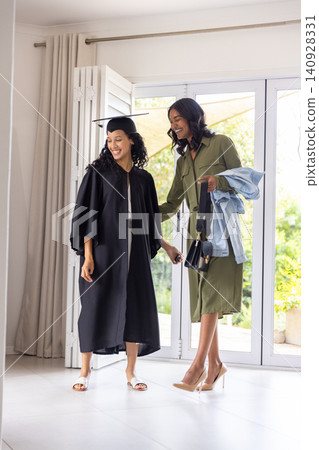 Mother guiding teenage daughter in home glass doorway holding graduation cap, gown Mother guiding teenage daughter in home glass doorway holding graduation cap, gown 140928331