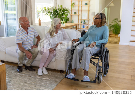 Senior friends chatting on sofa next to manual wheelchair in living room and plants, copy space Senior friends chatting on sofa next to manual wheelchair in living room and plants, copy space 140928335
